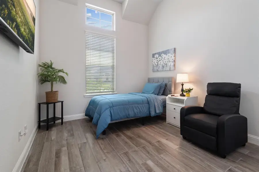 This cozy bedroom features a tall window for natural light, a comfortable bed with blue bedding, a white nightstand with a lamp, and a black recliner. The room has wood-look flooring and is accented with a plant and artwork for a serene atmosphere. This cozy bedroom features a tall window for natural light, a comfortable bed with blue bedding, a white nightstand with a lamp, and a black recliner. The room has wood-look flooring and is accented with a plant and artwork for a serene atmosphere.