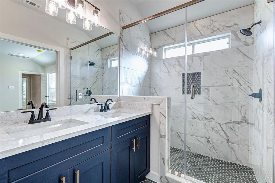 This luxurious primary bathroom showcases a spa-like experience with a stunning glass-enclosed walk-in shower adorned with exquisite marble tile and modern hexagon floor detailing. Dual vanities with navy cabinets and quartz countertops, paired with matte black fixtures, offer both elegance and practicality.