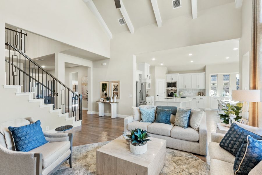 Open-concept living room with vaulted beamed ceiling, staircase, and white kitchen visible in background Open-concept living room with vaulted beamed ceiling, staircase, and white kitchen visible in background