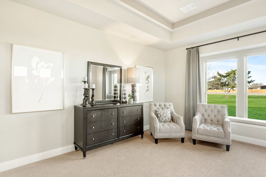 Bedroom sitting area with dark wood dresser, two tufted chairs, tray ceiling, and large window overlooking green lawn Bedroom sitting area with dark wood dresser, two tufted chairs, tray ceiling, and large window overlooking green lawn