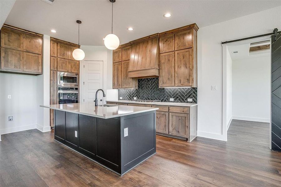 Kitchen featuring stainless steel appliances, hanging light fixtures, decorative backsplash, an island with sink, and dark wood-type flooring