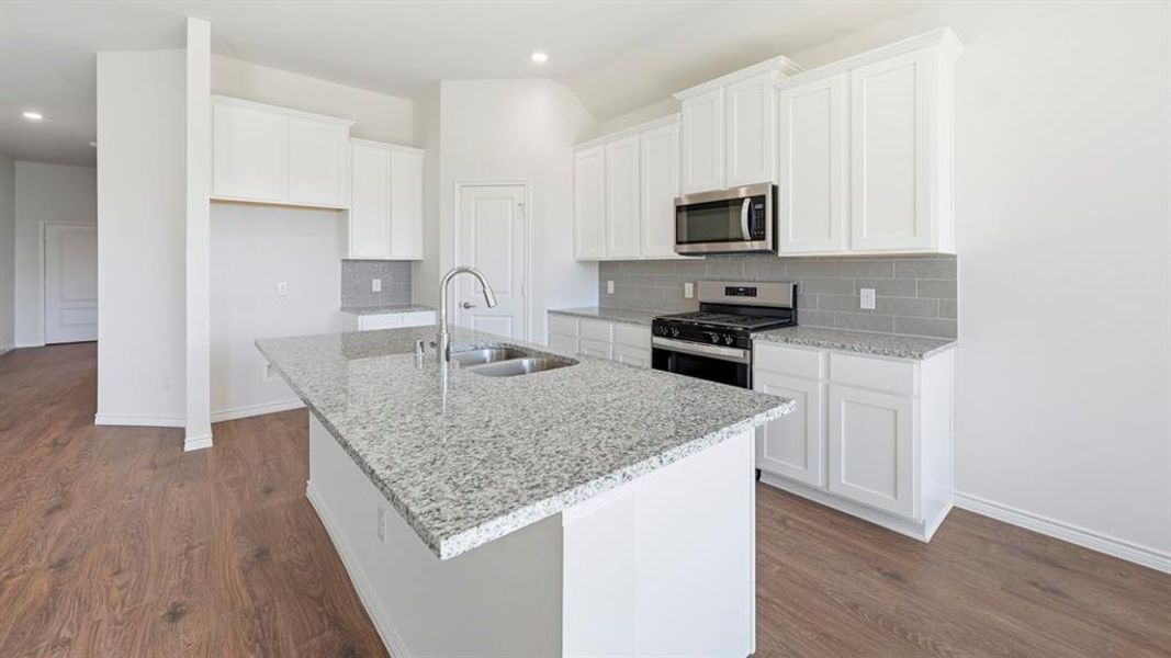 Open-concept kitchen featuring an island with a stainless steel sink, wood-finish flooring, white cabinetry, granite countertops, and stainless steel appliances including an oven range and microwave Open-concept kitchen featuring an island with a stainless steel sink, wood-finish flooring, white cabinetry, granite countertops, and stainless steel appliances including an oven range and microwave