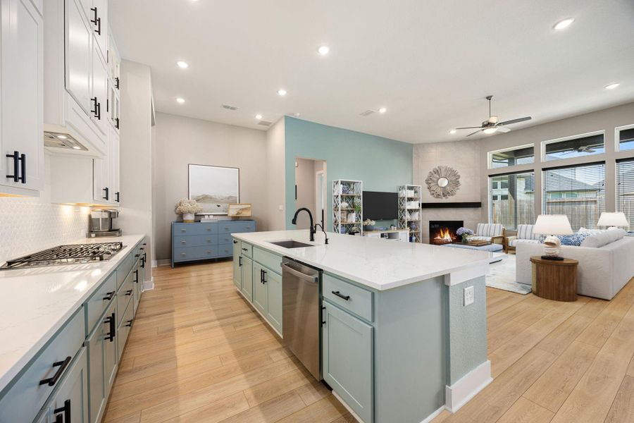 This kitchen and living area showcases an open-concept design with sleek quartz countertops, custom two-tone cabinetry, and a spacious island with a built-in sink. Recessed lighting and expansive windows brighten the space, complemented by warm wood-look flooring. The layout effortlessly connects the kitchen to the living area, where a modern fireplace and soaring ceilings create a comfortable yet refined setting.