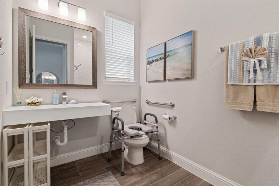 This ADA accessible bathroom features a modern design with a large mirror, sleek white vanity, and wood-look tile flooring. It includes accessible features like grab bars and an elevated toilet seat. Decor elements include calming beach-themed artwork and soft lighting. This ADA accessible bathroom features a modern design with a large mirror, sleek white vanity, and wood-look tile flooring. It includes accessible features like grab bars and an elevated toilet seat. Decor elements include calming beach-themed artwork and soft lighting.