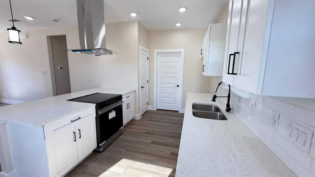 Modern kitchen featuring white shaker cabinetry, speckled countertops, wood-finish flooring, a stainless steel range hood, and a dual basin sink with a matte black faucet Modern kitchen featuring white shaker cabinetry, speckled countertops, wood-finish flooring, a stainless steel range hood, and a dual basin sink with a matte black faucet
