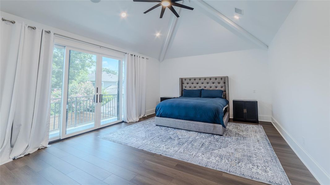 This spacious bedroom features vaulted ceilings, hardwood floors, and a large sliding glass door leading to a balcony. The room is filled with natural light and has a modern, elegant design with a plush bed and large area rug.
