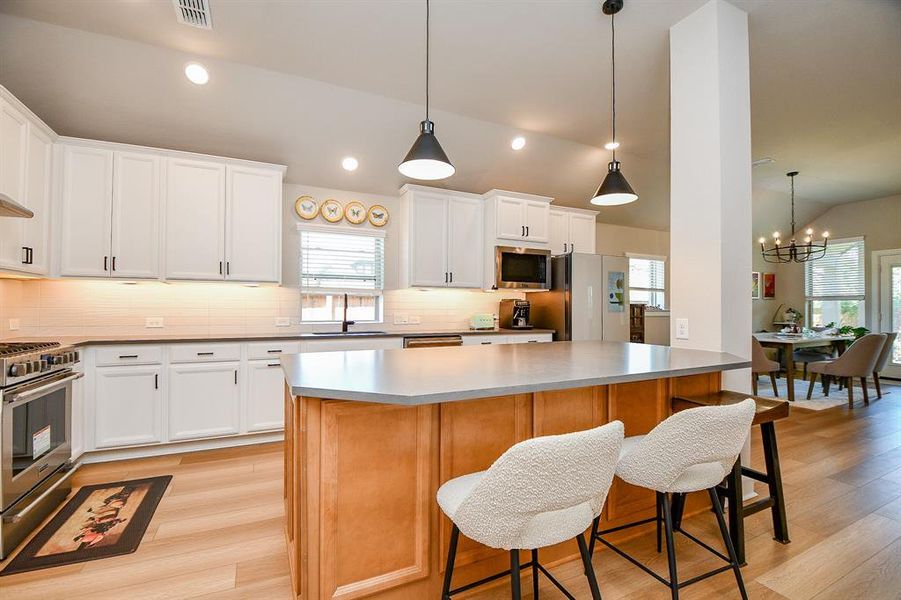 This modern kitchen features white cabinetry, stainless steel appliances, and a spacious island with seating. It's well-lit with pendant lights and opens into a dining area with a stylish chandelier, creating a warm and inviting space.