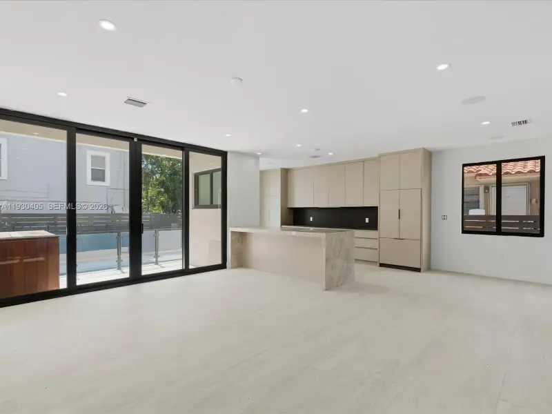 Open-concept interior featuring light-toned flooring, custom flat-panel cabinetry, a stone-finished kitchen island, integrated appliances, and floor-to-ceiling sliding glass doors Open-concept interior featuring light-toned flooring, custom flat-panel cabinetry, a stone-finished kitchen island, integrated appliances, and floor-to-ceiling sliding glass doors
