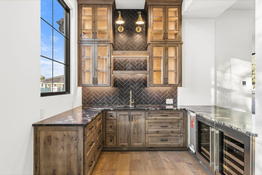 Indoor wet bar with tasteful backsplash, light wood-style flooring, dark stone counters, wine cooler, and glass insert cabinets