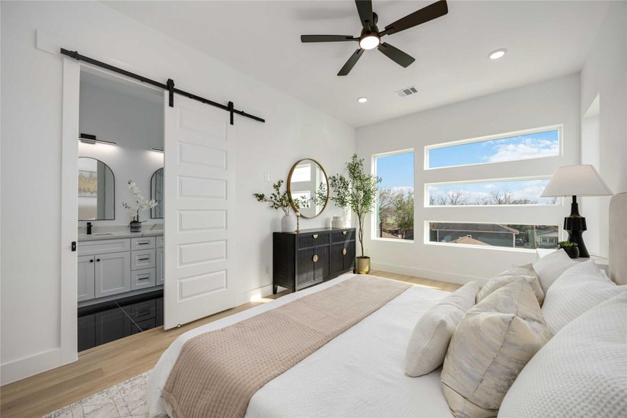 This stunning primary suite showcases expansive modern windows that frame serene outdoor views and fill the space with natural light, complemented by warm wood-look flooring, soft neutral décor, and a sleek ceiling fan; a stylish sliding barn door opens to the luxurious en-suite bathroom, enhancing the room’s seamless and contemporary design.