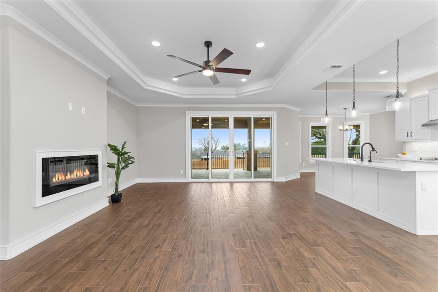 Unfurnished living room featuring a raised ceiling, a ceiling fan, a glass covered fireplace, dark wood-style floors, and ornamental molding Unfurnished living room featuring a raised ceiling, a ceiling fan, a glass covered fireplace, dark wood-style floors, and ornamental molding