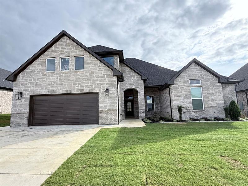 French country inspired facade with a front lawn, driveway, roof with shingles, brick siding, and a garage