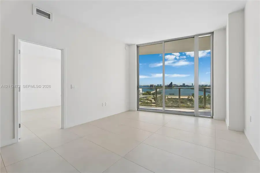 Floor-to-ceiling glass sliding doors, expansive tile flooring, balcony with metal railing, and panoramic water and urban skyline backdrop