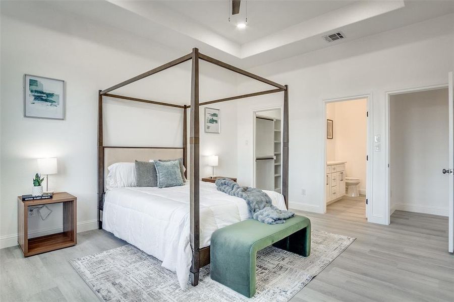 Bedroom featuring light wood-style flooring, a ceiling fan, a spacious closet, a tray ceiling, and ensuite bathroom Bedroom featuring light wood-style flooring, a ceiling fan, a spacious closet, a tray ceiling, and ensuite bathroom