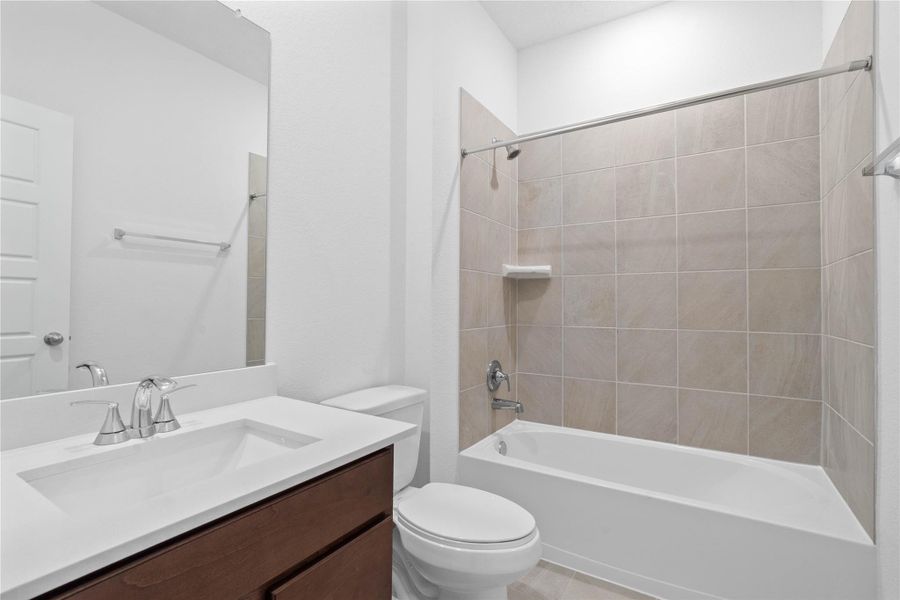 The secondary bathroom offers tile flooring, a bath/shower combination with a stylish tile surround, rich stained wood cabinetry and elegant light countertops, a mirror, and sleek fixtures with modern finishes.