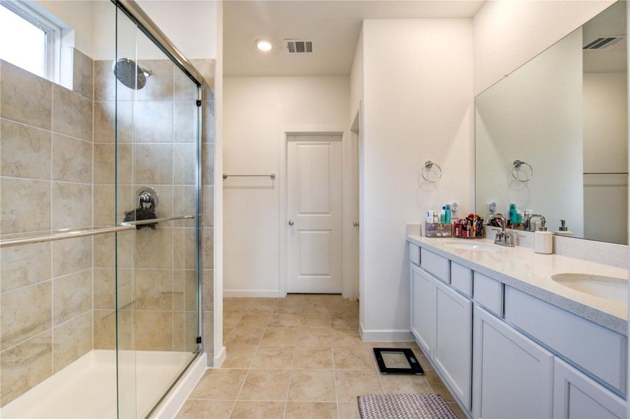 Primary Bathroom features elevated ceilings with recessed lighting and a calming neutral palette, creating a bright and serene atmosphere. It is appointed with double vanities offering generous counter space and storage, along with a sleek glass-enclosed shower. Thoughtfully designed, this space provides a refined setting for both comfort and everyday functionality.