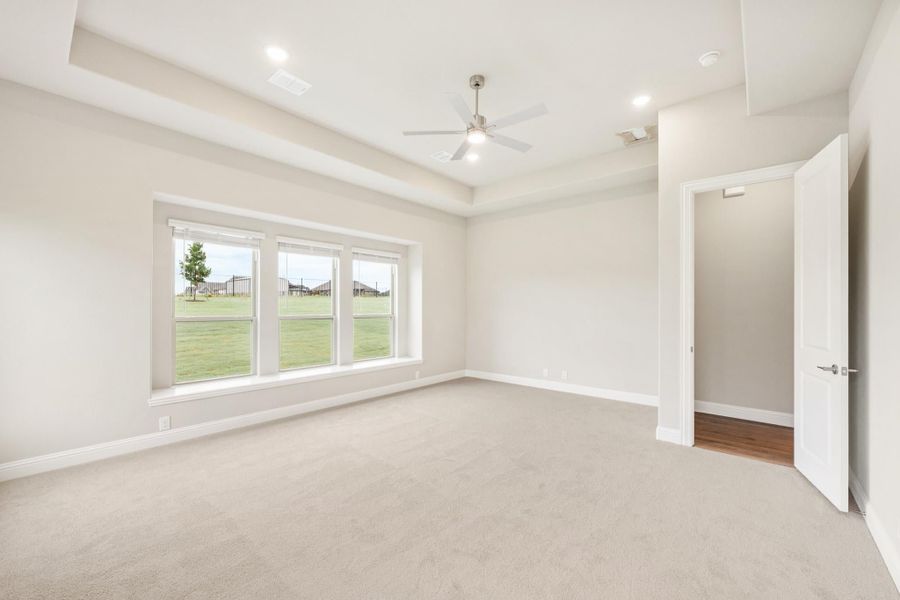 Empty bedroom with tray ceiling, ceiling fan, carpet flooring, and triple window overlooking backyard