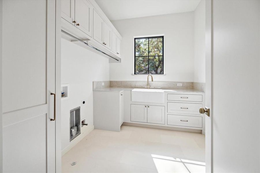 This full size laundry room is both functional and stylish, featuring crisp white cabinetry with brass hardware for a touch of elegance. A large farmhouse sink sits beneath a black-framed window, bringing in natural light. With overhead cabinets offering plenty of storage, there is also ample space for a washer and dryer side by side with both electric and gas dryer connections.