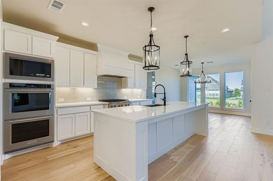 Kitchen with backsplash, stainless steel appliances, white cabinets, an island with sink, and hanging light fixtures