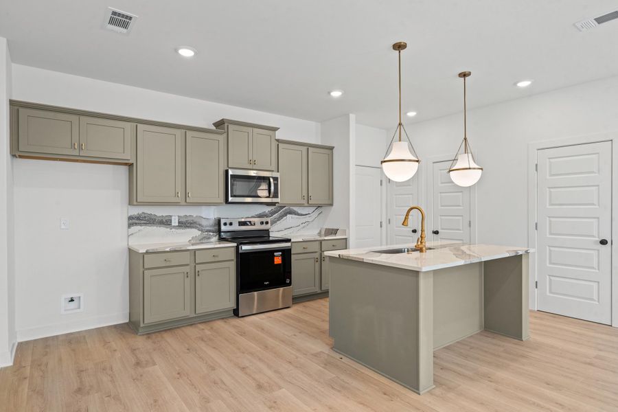 Kitchen featuring light wood-finish flooring, island with a built-in sink and gold-tone faucet, two pendant lights, recessed lighting, and gray cabinetry with stainless steel appliances