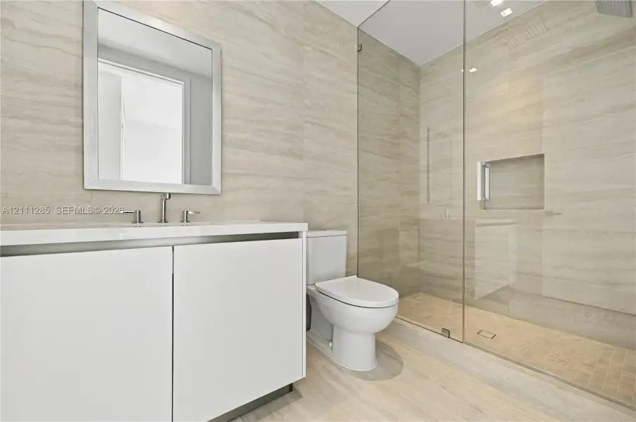 Minimalist vanity featuring a smooth-finish white cabinet and rectangular sink with metal fixtures, complemented by wall-to-wall linear-patterned tile, a seamless glass-enclosed shower with a built-in recessed niche, and mosaic floor tiling Minimalist vanity featuring a smooth-finish white cabinet and rectangular sink with metal fixtures, complemented by wall-to-wall linear-patterned tile, a seamless glass-enclosed shower with a built-in recessed niche, and mosaic floor tiling