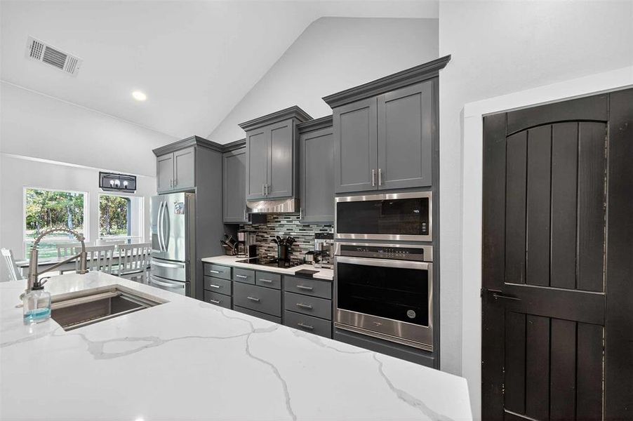 Kitchen featuring decorative backsplash, appliances with stainless steel finishes, light stone counters, gray cabinetry, and high vaulted ceiling Kitchen featuring decorative backsplash, appliances with stainless steel finishes, light stone counters, gray cabinetry, and high vaulted ceiling