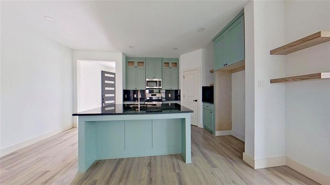 Kitchen featuring green cabinets, an island with sink, light wood-style floors, stainless steel appliances, and dark stone counters