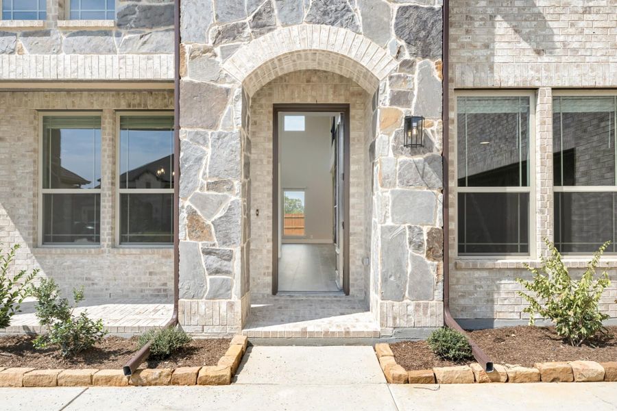 Home entrance with stone arch surround, open front door, brick exterior, and small landscaped garden beds on each side.