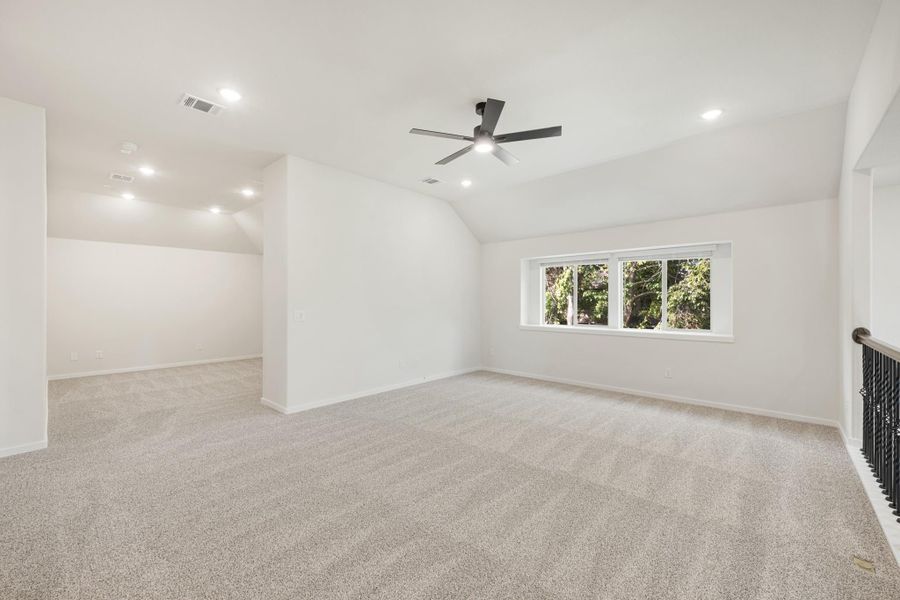 Spacious empty room with vaulted ceiling, ceiling fan, carpet flooring, and large window near staircase railing