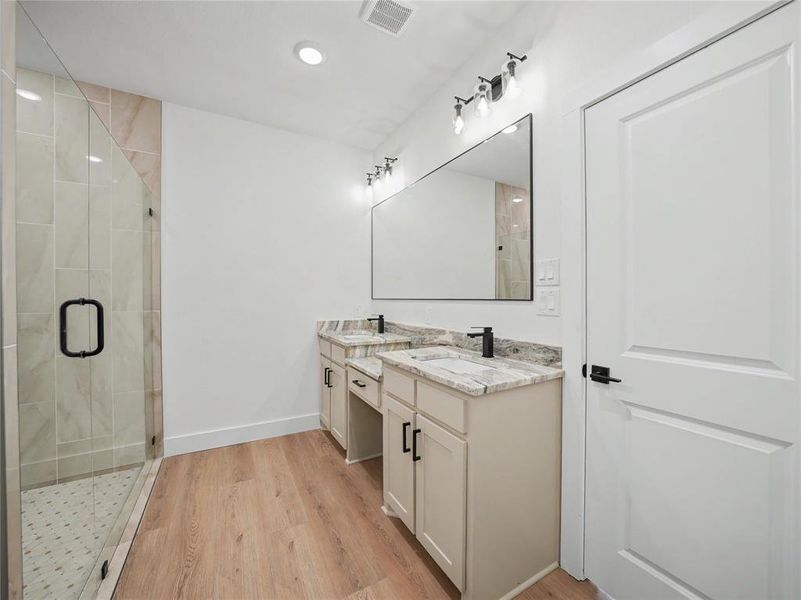 Bathroom featuring a frameless glass shower enclosure with tiled walls and a mosaic floor, a dual vanity with light-toned cabinetry, granite countertops, and matte black fixtures, wood-finish flooring, and recessed lighting Bathroom featuring a frameless glass shower enclosure with tiled walls and a mosaic floor, a dual vanity with light-toned cabinetry, granite countertops, and matte black fixtures, wood-finish flooring, and recessed lighting