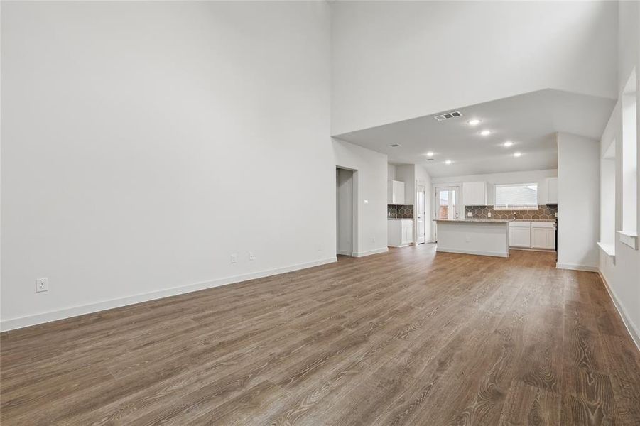 Unfurnished living room featuring a high ceiling, wood finished floors, baseboards, and visible vents Unfurnished living room featuring a high ceiling, wood finished floors, baseboards, and visible vents