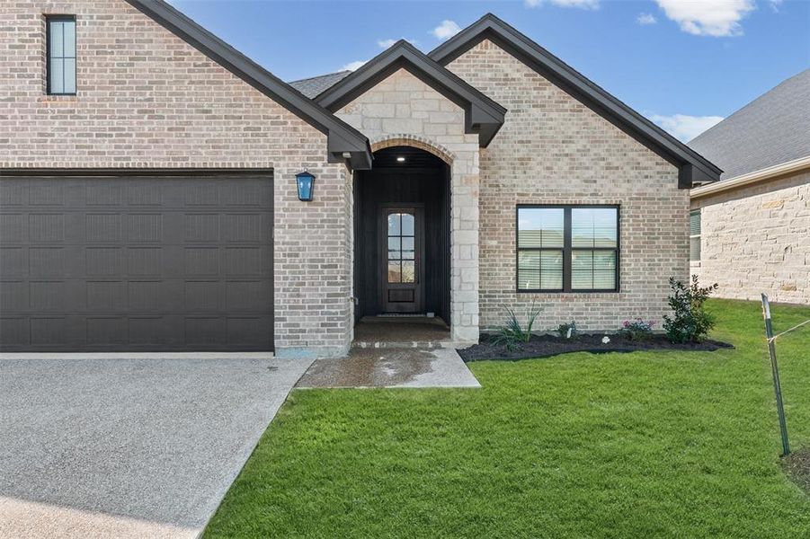 View of exterior entry with a lawn, brick siding, driveway, a garage, and stone siding