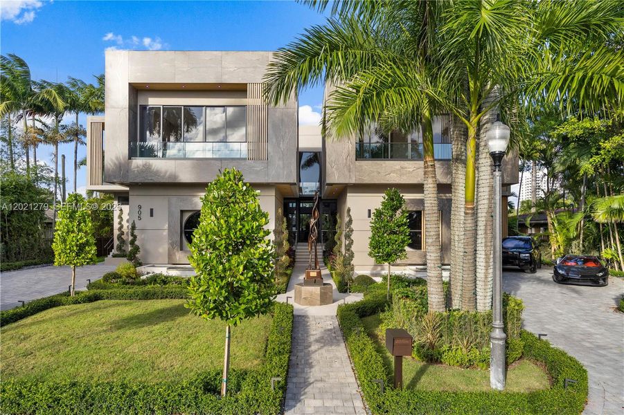 Two-story modern luxury home with stone façade, paver walkway, bronze sculpture, manicured hedges, tropical landscaping, second-level glass balcony, and wide motor court.