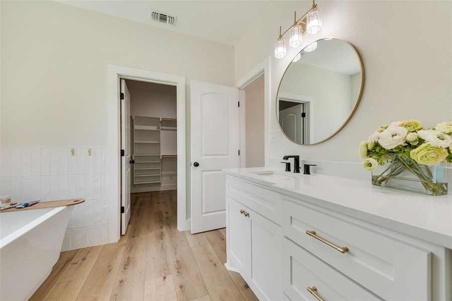 Bathroom featuring a white freestanding bathtub with vertical subway tile wainscoting, a white vanity with a rectangular undermount sink, a large gold-framed oval mirror, and light wood-finish flooring