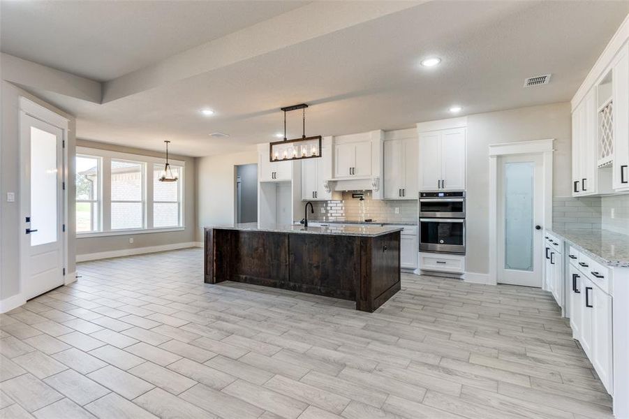 Kitchen with light stone countertops, stainless steel double oven, a center island with sink, and white cabinetry