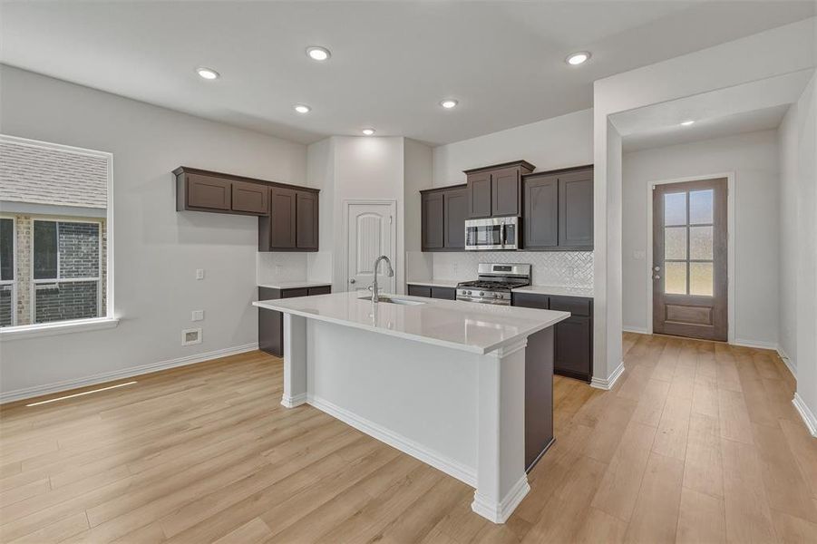 Contemporary kitchen featuring a large central island with a sink, dark wood cabinetry, stainless steel appliances, and wood-finish flooring Contemporary kitchen featuring a large central island with a sink, dark wood cabinetry, stainless steel appliances, and wood-finish flooring