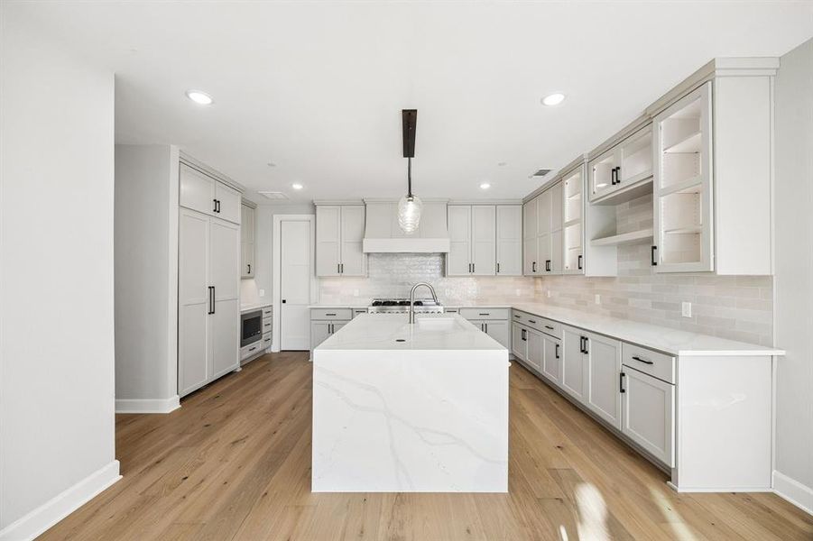 Kitchen featuring hanging light fixtures, open shelves, light stone countertops, a center island with sink, and light wood-style flooring Kitchen featuring hanging light fixtures, open shelves, light stone countertops, a center island with sink, and light wood-style flooring