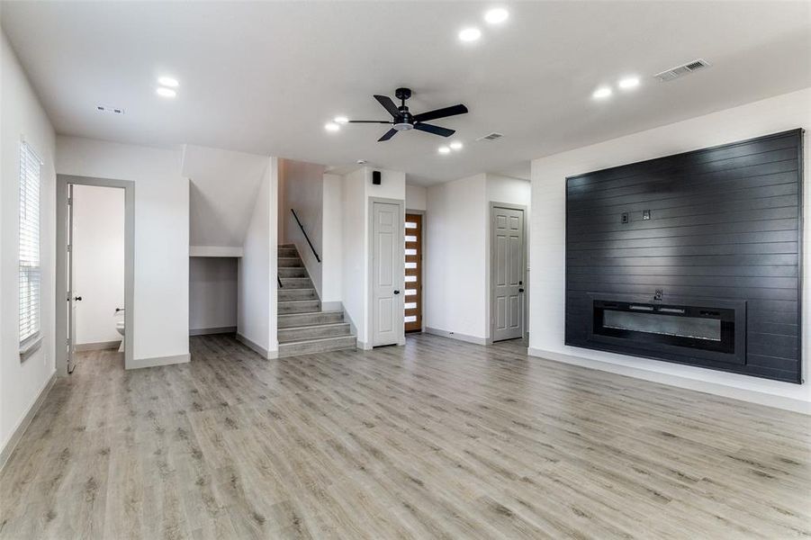Unfurnished living room with ceiling fan, a glass covered fireplace, light wood-style flooring, stairway, and recessed lighting Unfurnished living room with ceiling fan, a glass covered fireplace, light wood-style flooring, stairway, and recessed lighting