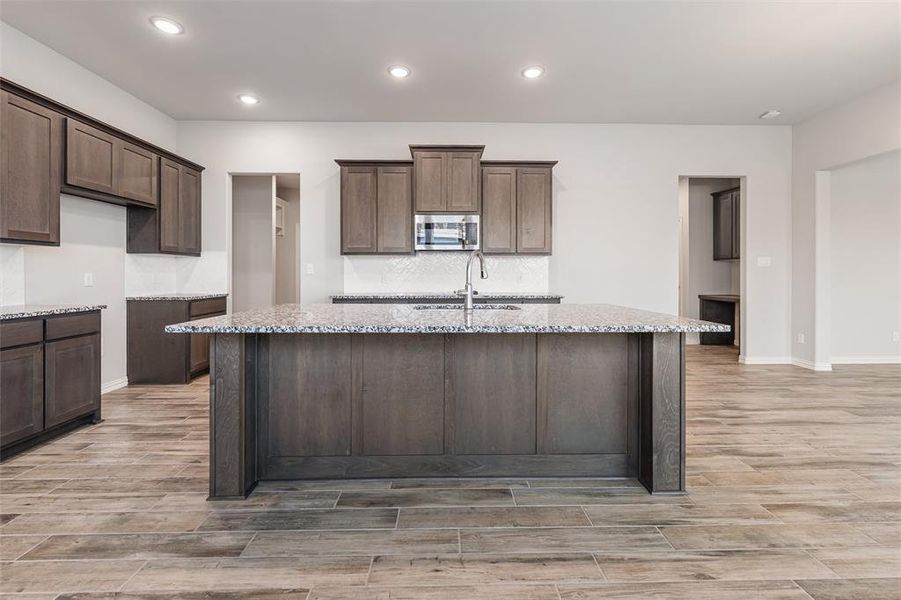 Kitchen featuring light stone counters, wood finish floors, an island with sink, recessed lighting, and dark brown cabinets Kitchen featuring light stone counters, wood finish floors, an island with sink, recessed lighting, and dark brown cabinets