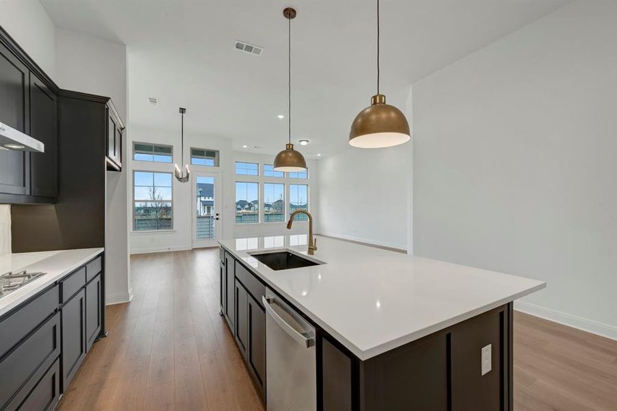 Kitchen featuring a kitchen island with sink, dishwasher, light wood-style floors, hanging light fixtures, and light stone counters