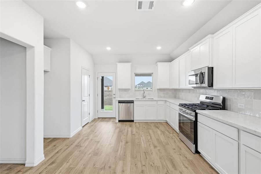 Kitchen featuring appliances with stainless steel finishes, white cabinetry, light wood-type flooring, decorative backsplash, and light stone countertops