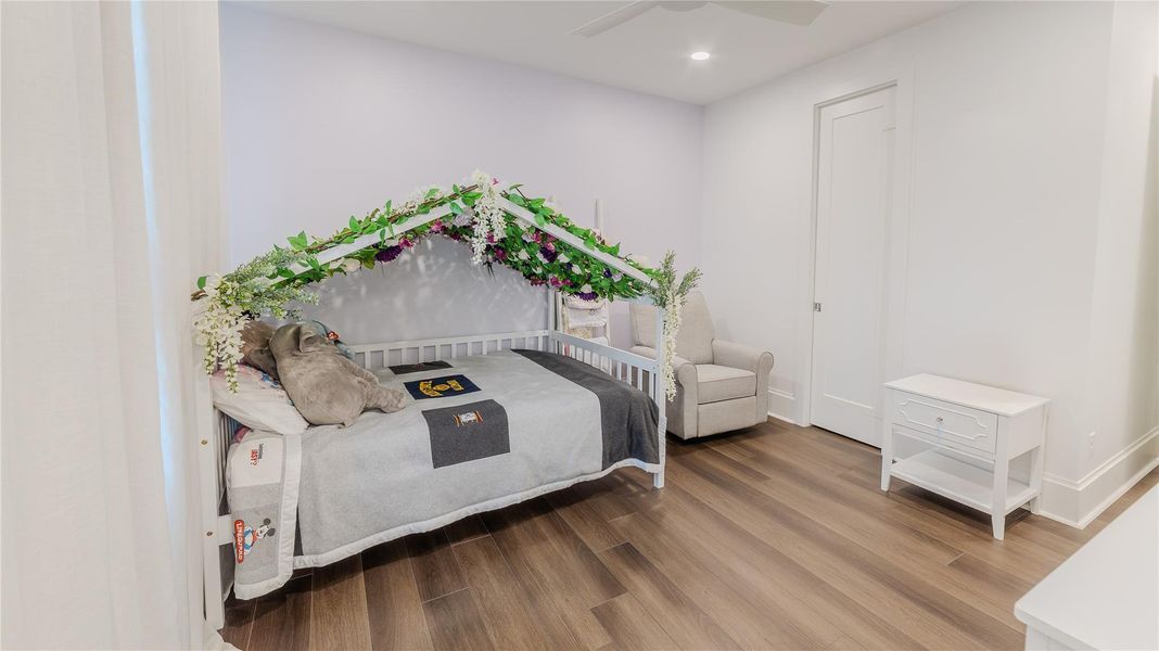This cozy children's bedroom features a charming bed adorned with a floral canopy, a plush stuffed animal, and a comfortable armchair. The room has warm wood flooring, soft lighting, and a small white nightstand, creating a welcoming and serene atmosphere.