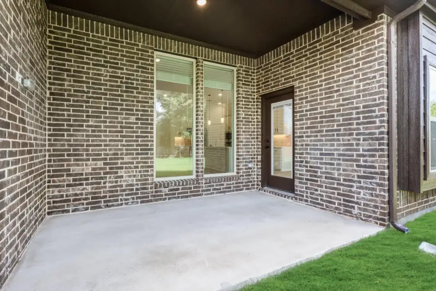 Covered back patio with concrete floor, dark brick walls, large window, and glass door leading inside.
