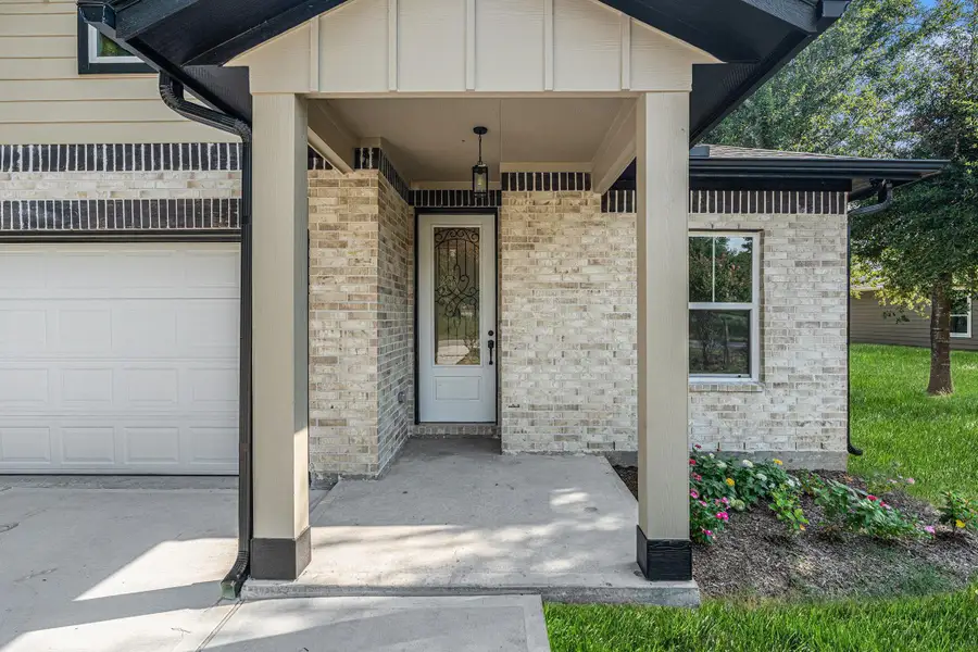 Charming entryway with a covered porch featuring light brickwork and a decorative front door. Adjacent to the garage, the area is complemented by a small landscaped garden and lush green lawn.