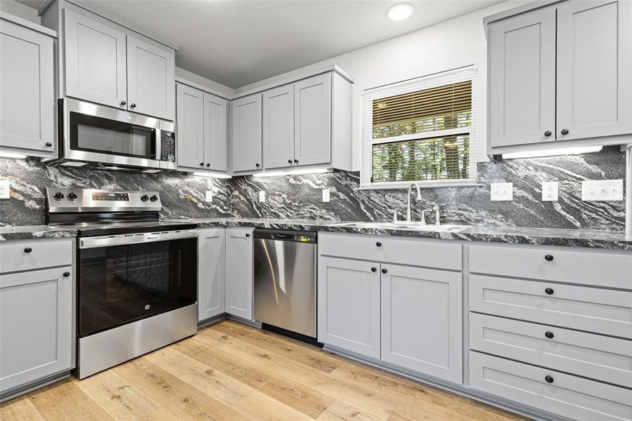 Kitchen featuring stainless steel appliances, backsplash, light wood-style flooring, dark stone counters, and gray cabinets Kitchen featuring stainless steel appliances, backsplash, light wood-style flooring, dark stone counters, and gray cabinets