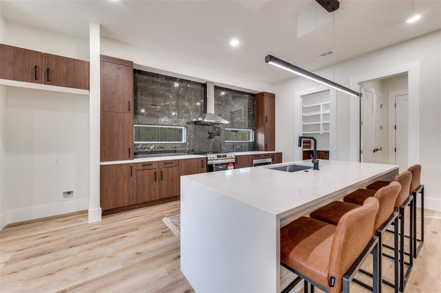 Modern kitchen featuring a large waterfall island with integrated sink, dark wood-finish cabinetry, light wood-finish flooring, and a striking dark stone-finish backsplash with horizontal windows