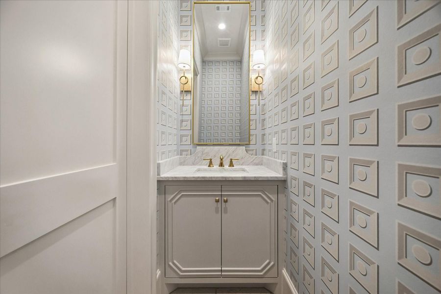 Elegant powder bath featuring a marble countertop and backsplash, complemented by a gorgeous custom vanity with polished hardware. Designer wallcoverings add striking dimension, while a framed mirror and beautiful sconces create a sophisticated, elevated ambiance.
