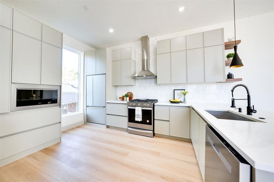 Kitchen with wall chimney exhaust hood, hanging light fixtures, stainless steel appliances, sink, and light hardwood / wood-style floors