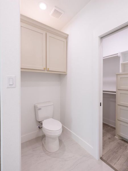 This photo shows a clean, modern bathroom with a white toilet and cabinetry above for storage. The room features a light, neutral color scheme and opens into a spacious closet area with shelving and drawers.