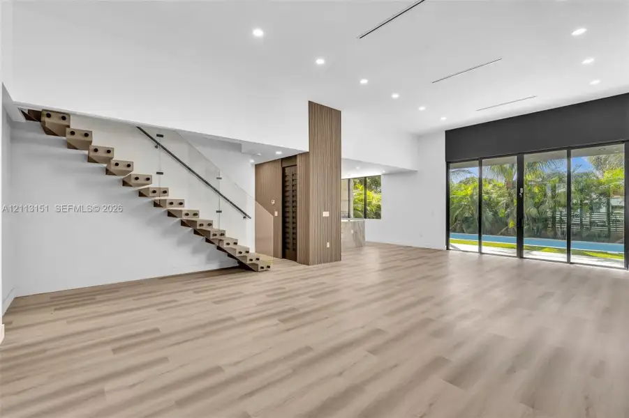 Open-concept living space featuring wood-finish flooring, floating staircase with glass railings, recessed ceiling lighting, and expansive floor-to-ceiling sliding glass doors Open-concept living space featuring wood-finish flooring, floating staircase with glass railings, recessed ceiling lighting, and expansive floor-to-ceiling sliding glass doors